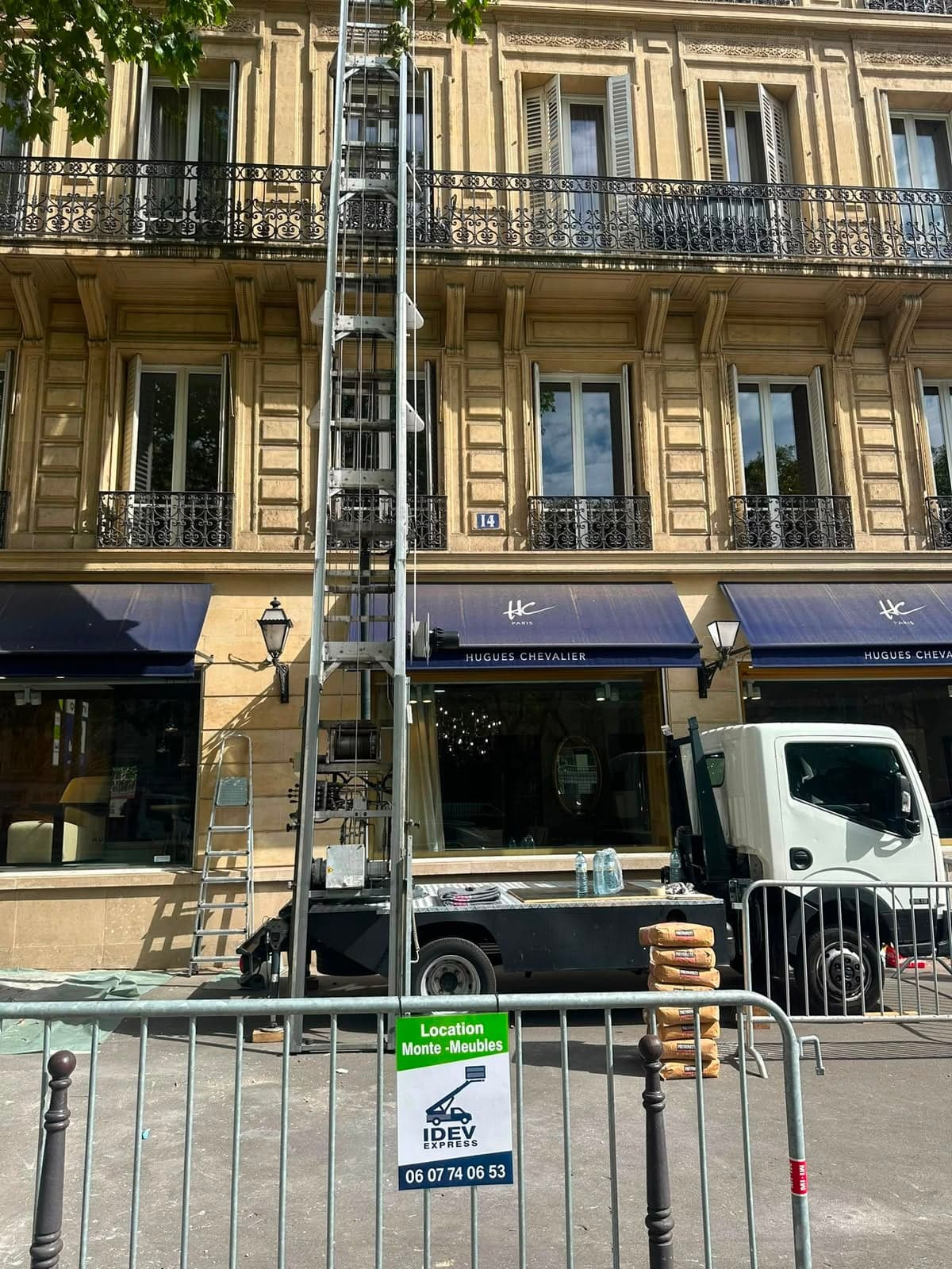 Monte-meuble IDEV EXPRESS dressé le long d’une façade haussmannienne parisienne, sacs de ciment empilés à son pied derrière les barrières de chantier